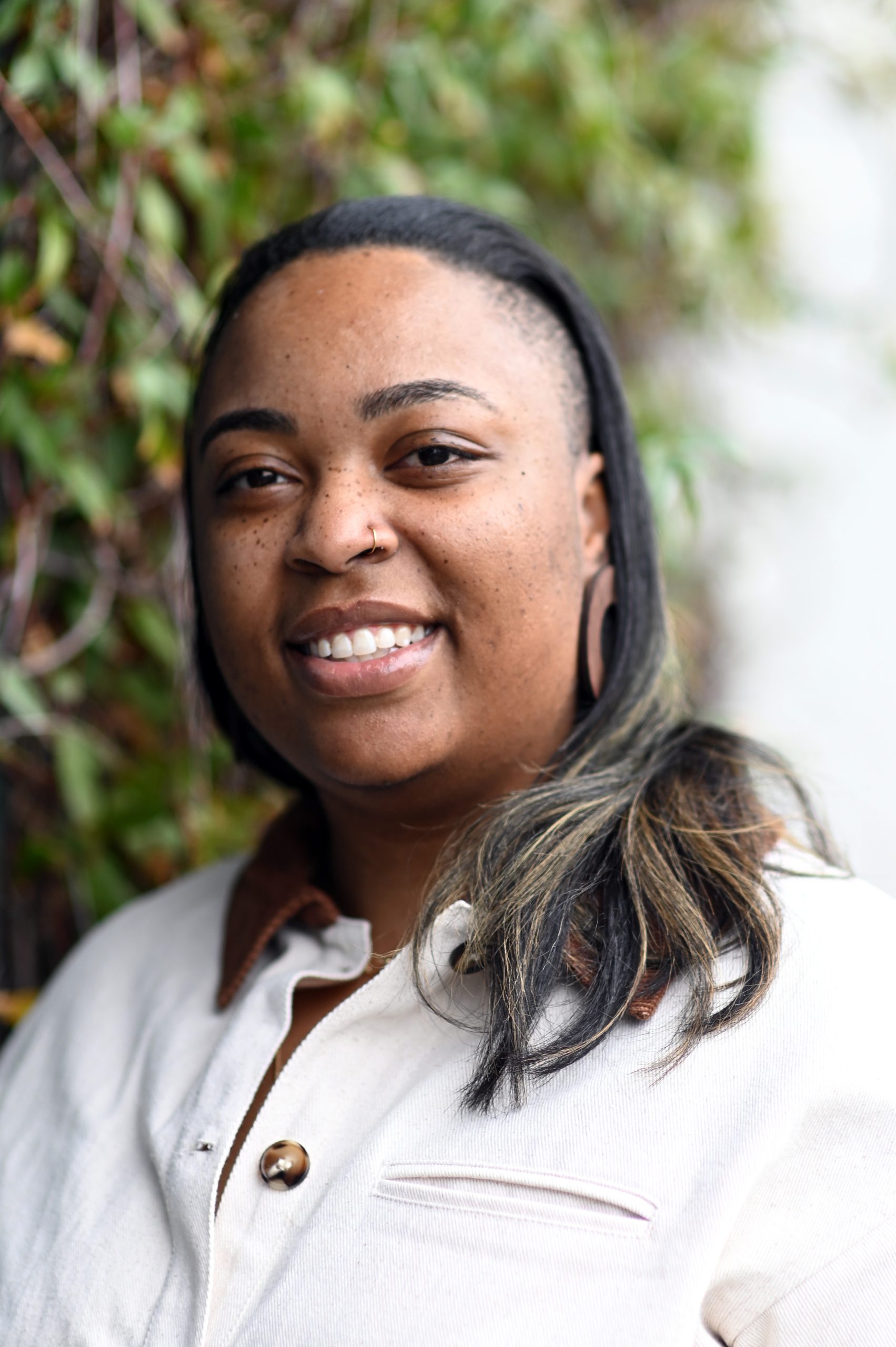 Headshot of BT Paschall. They are smiling broadly, facing the camera, angled slightly to the left. She has glowing brown skin and straight shoulder-length, black hair. Beloved is wearing a cream colored button up shirt. There is a green bush behind them. 